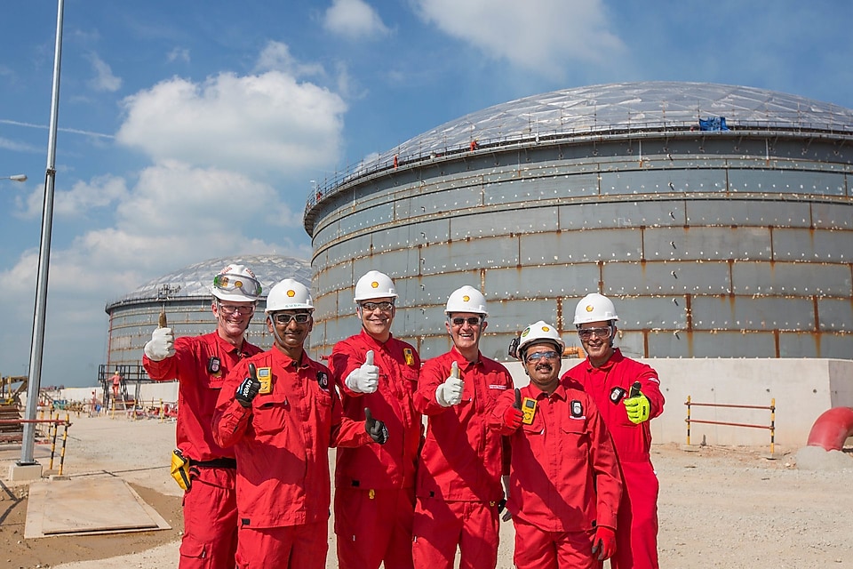 six workers showing thumbs up with two large crude oil tanks in their background