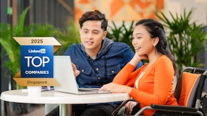 Two individuals are seated at a round table, engaged in a discussion while looking at a laptop. The person on the left is wearing a blue jacket, and the person on the right is wearing an orange cardigan. In the foreground, there is an award plaque that reads "2025 LinkedIn Top Companies Singapore." The background features indoor plants and blurred decor elements.