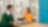 A man in a green sweater and a woman in an orange top are sitting at a table in an office. The woman is holding a tablet, and there is a yellow coffee cup on the table next to her. A blue folder is in front of the man. Large windows in the background reveal an urban view.