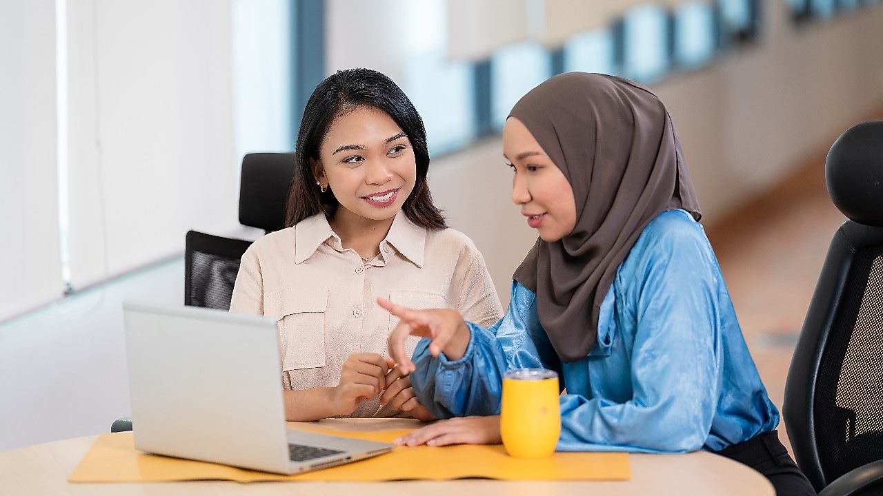Two individuals are seated at a table in an office setting, engaged in a discussion. One individual is wearing a light-colored shirt and the other is wearing a blue top with a headscarf. They are looking at a laptop screen, and one of them is pointing towards it. A yellow cup is placed on the table next to the laptop.