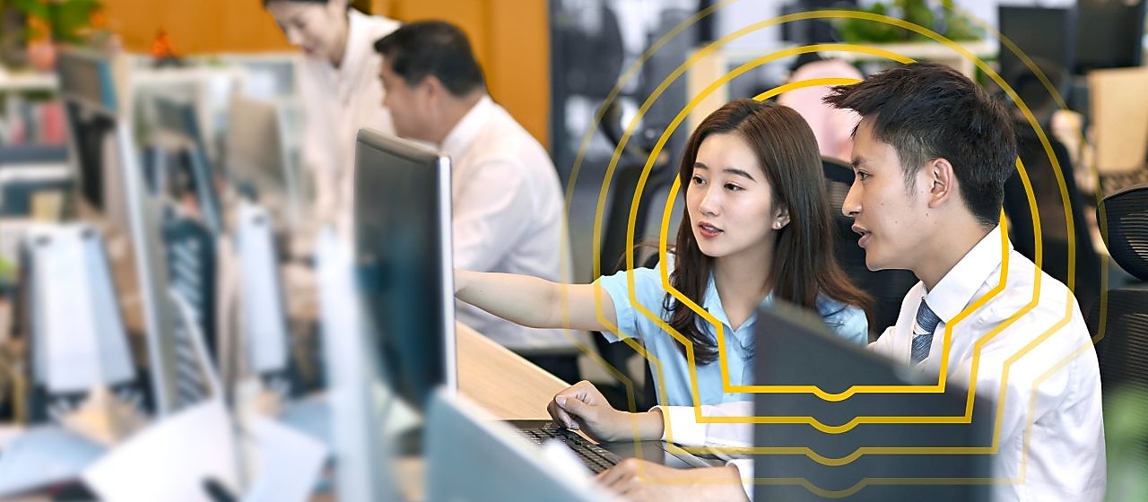 Collaborating employees working at computer desks in a modern office with multiple monitors and green plants in the background.