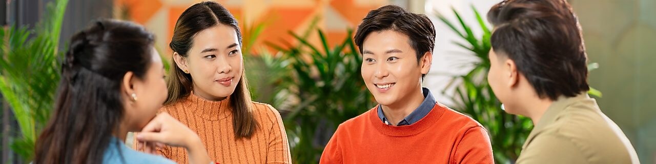 Four people sitting around a round table in a modern office, engaged in discussion with notebooks and mugs on the table.