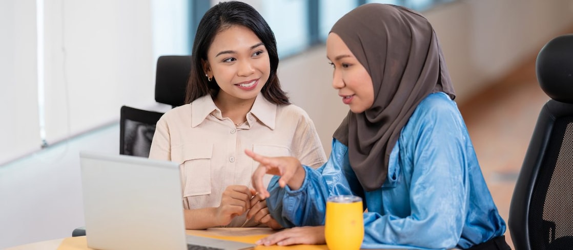 Two individuals are seated at a table in an office setting, engaged in a discussion. One individual is wearing a light-colored shirt and the other is wearing a blue top with a headscarf. They are looking at a laptop screen, and one of them is pointing towards it. A yellow cup is placed on the table next to the laptop.