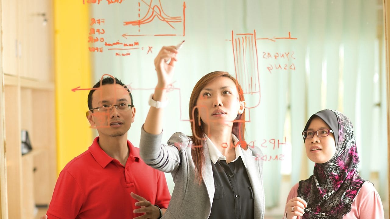 Colleagues gathered standing and discussing metrics on a glass board