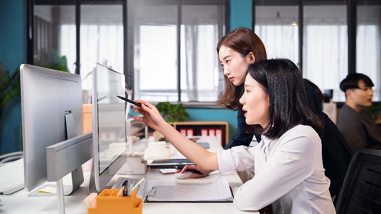 Two colleagues collaborating at a desk, reviewing content on a computer monitor with documents and office supplies nearby.