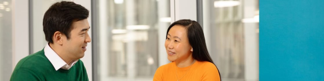 A man in a green sweater and a woman in an orange top are sitting at a table in an office. The woman is holding a tablet, and there is a yellow coffee cup on the table next to her. A blue folder is in front of the man. Large windows in the background reveal an urban view.