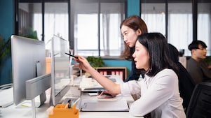 Two colleagues collaborating at a desk, reviewing content on a computer monitor with documents and office supplies nearby.