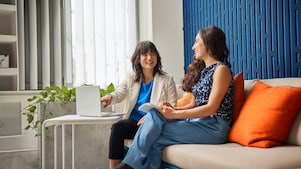 Two people sitting on a modern sofa with orange cushions, discussing something near a laptop in a bright, stylish room.