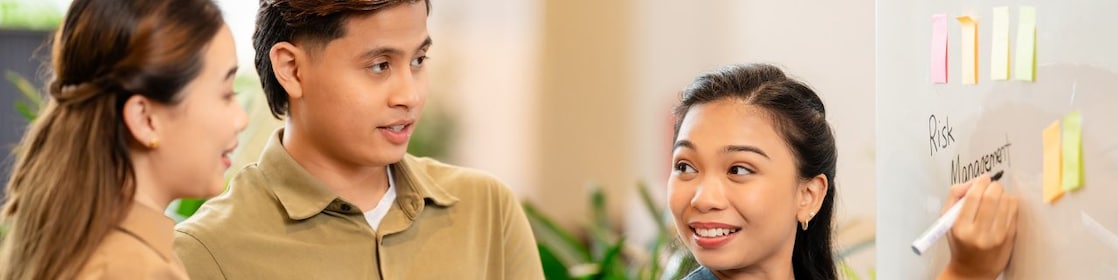 Three people collaborating at a whiteboard with sticky notes, discussing risk management and taking notes in a notebook.
