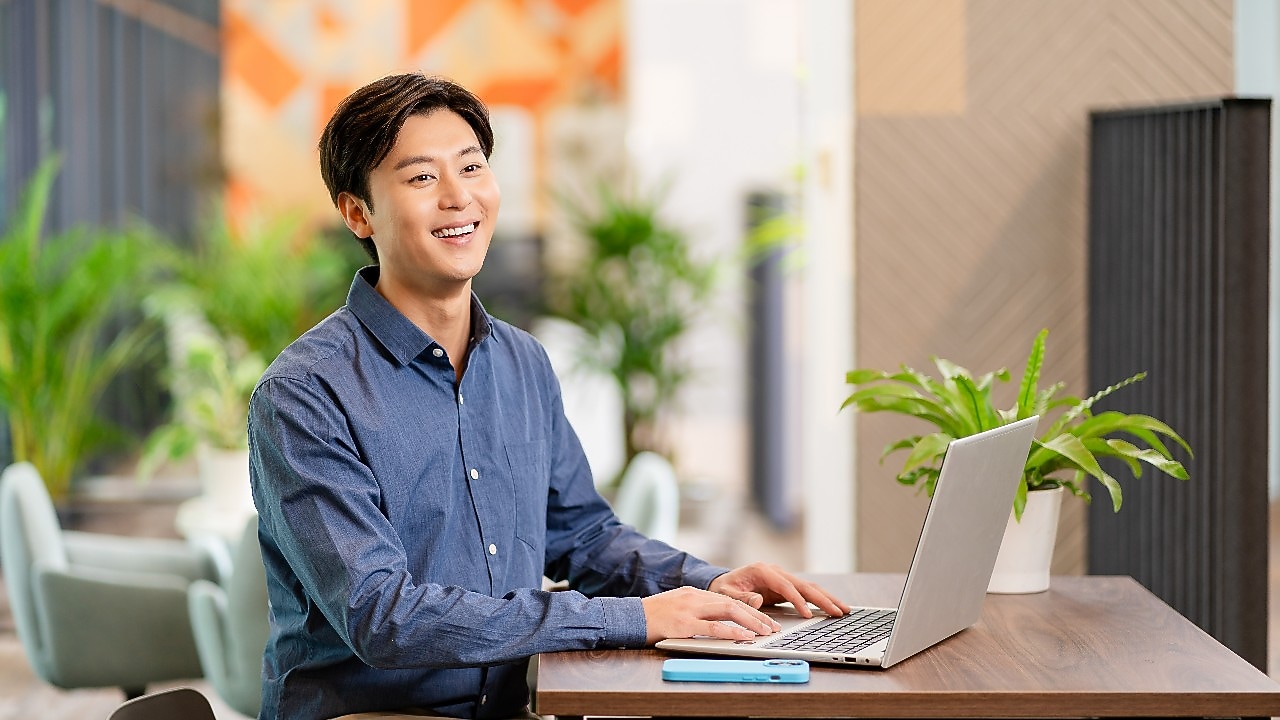 Person seated at a wooden table using a laptop, with a smartphone and potted plants in a bright modern workspace.