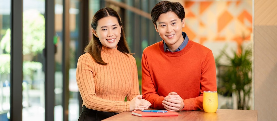 Two people in orange sweaters standing at a high table with a notebook, smartphone, and yellow mug in a bright modern office.