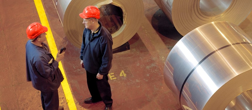 Two factory workers standing next to rolls of steel